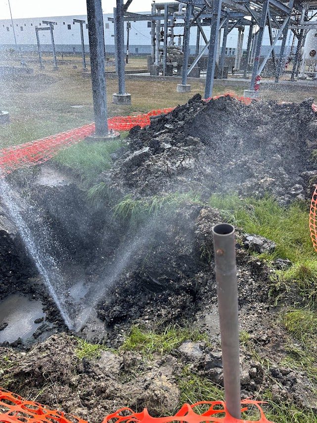 Water spraying from a broken line at an industrial facility behind a safety fence