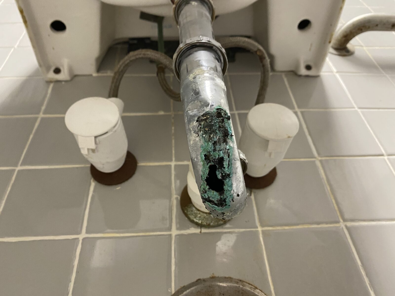 Corroded chrome tailpiece with a hole through it under a commercial lavatory