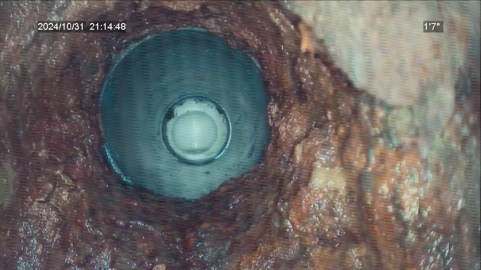 Sewer camera view down a corroded pipe toward a clean fitting further along the line